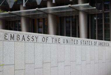 The front of a US Government building with a grey stone wall out front with Embassy of the United States of America engraved at the top.